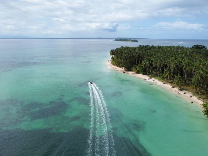 A small boat navigates through turquoise waters, framed by palm trees and pristine white sand beaches on Zapatilla Island, Panama.
